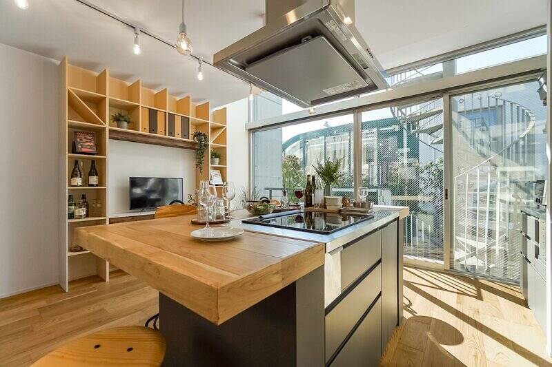 3口のIHヒーターを備えたキッチンが空間の主役。ダイニングバー風にテーブルと一体化しており、手を動かしながら団らんが可能です。壁面には、テレビボードを兼ねたオープンシェルフが造作されています。